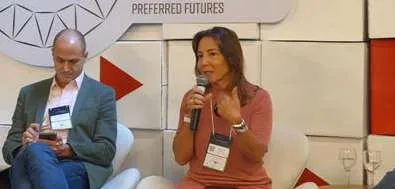 Fernanda Camargo falando ao microfone durante evento enquanto homem ao lado consulta celular.