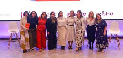 Dez mulheres em pé, sorrindo, alinhadas em um espaço interno com cadeiras brancas ao fundo.