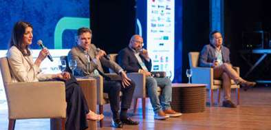 Quatro pessoas sentadas em cadeiras no palco participando de um painel de discussão em evento.