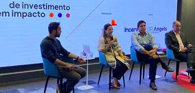 Quatro pessoas sentadas em cadeiras azuis em um painel de discussão com fundo com texto sobre investimento de impacto.