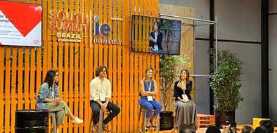 Quatro pessoas sentadas em cadeiras durante um painel em um evento com parede de madeira atrás exibindo os logotipos do South Summit Brazil e IE University.
