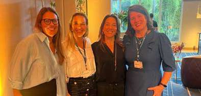 Quatro mulheres sorrindo, posando juntas em ambiente interno com iluminação quente.