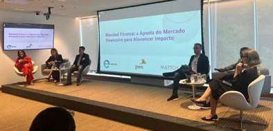 Mesa-redonda com cinco pessoas sentadas discutindo em evento com telas projetadas ao fundo sobre finanças e mercado financeiro.