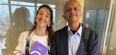 Dois adultos sorridentes, uma mulher segurando um documento roxo, em ambiente interno com parede de vidro e persianas.