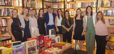 Dez pessoas em pé sorrindo dentro de uma livraria, com prateleiras cheias de livros ao fundo e uma mesa com livros na frente.