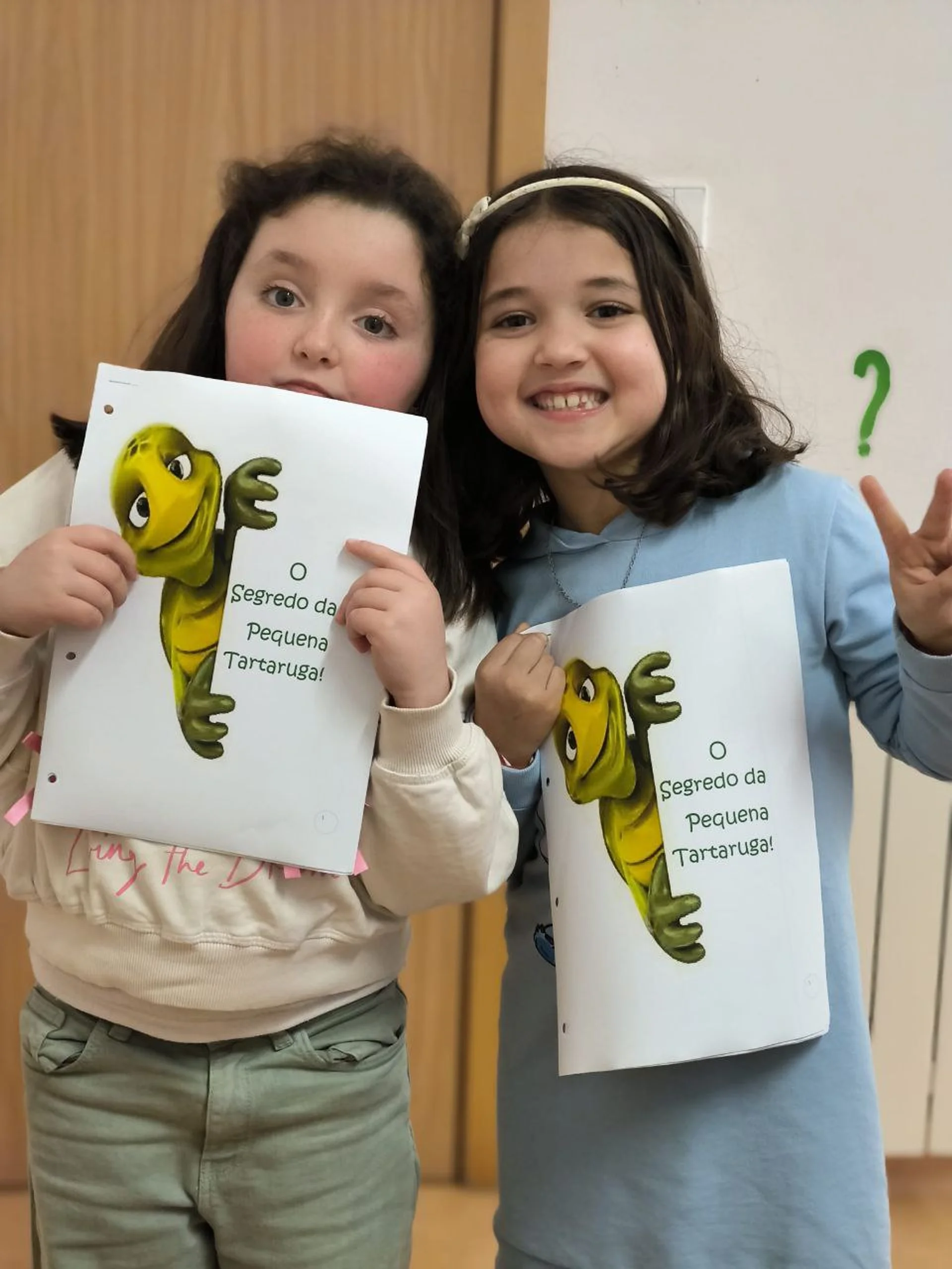 Duas meninas seguram livros com a capa mostrando uma tartaruga verde e o título 'O Segredo da Pequena Tartaruga'.
