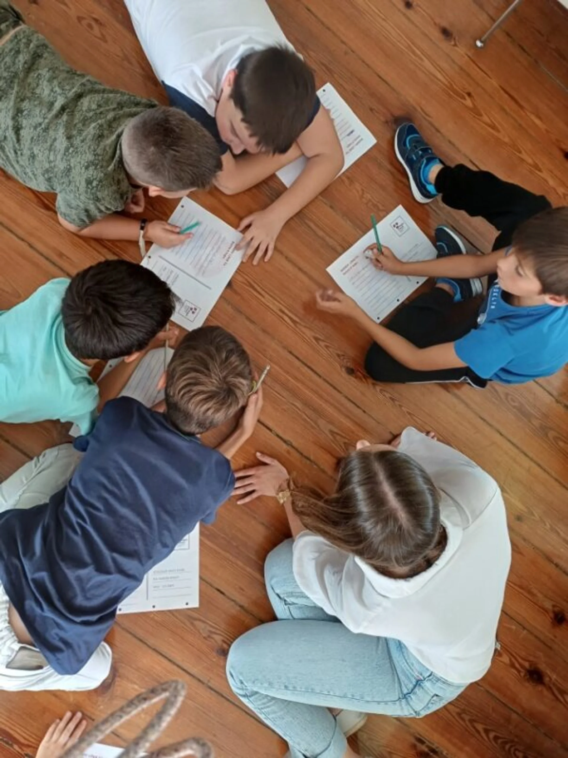 Grupo de cinco crianças e uma jovem sentados e deitados no chão de madeira a trabalhar em fichas de papel com lápis.