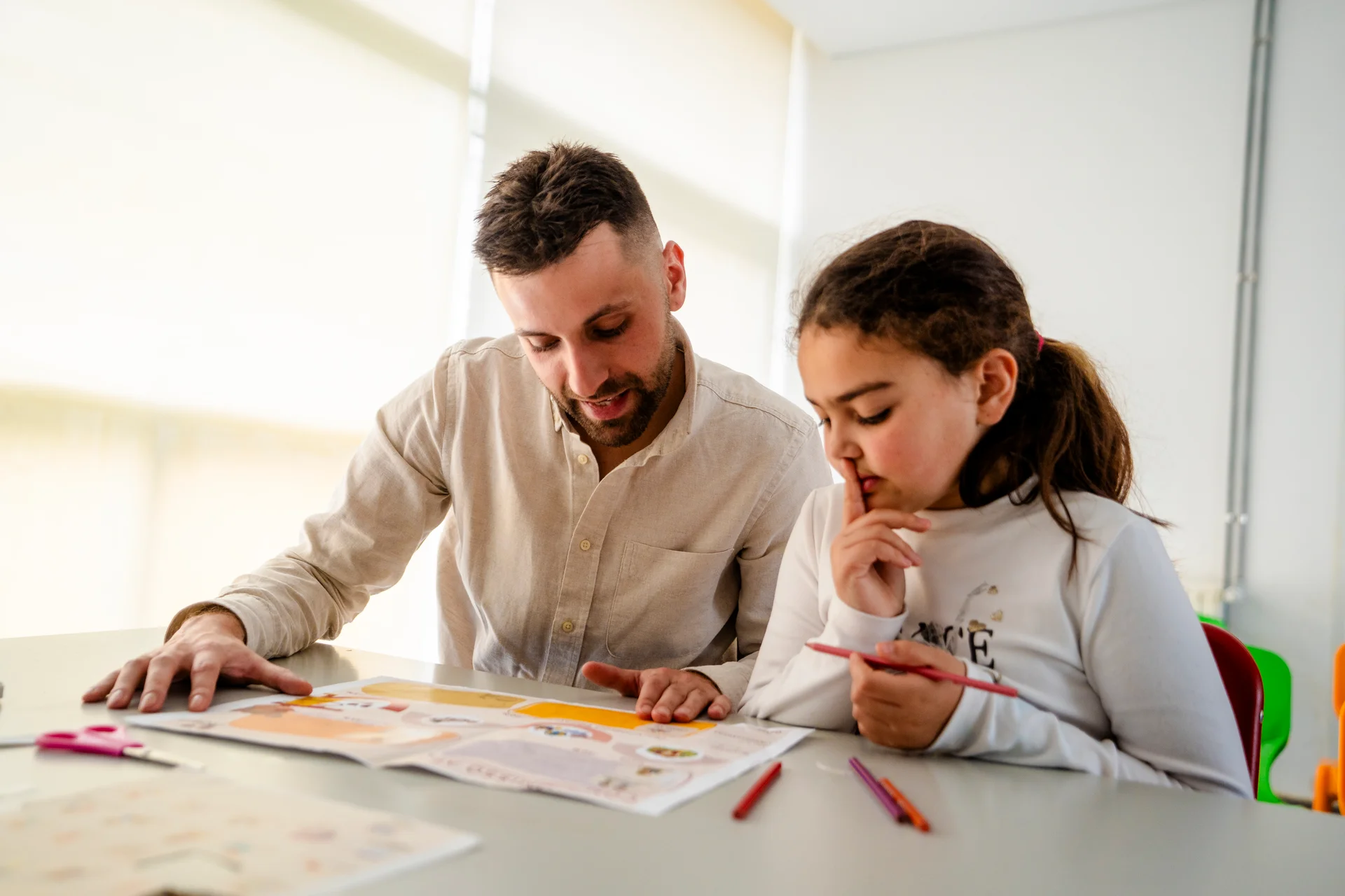 Homem ajudando uma menina a ler um livro em uma mesa com lápis de cor.