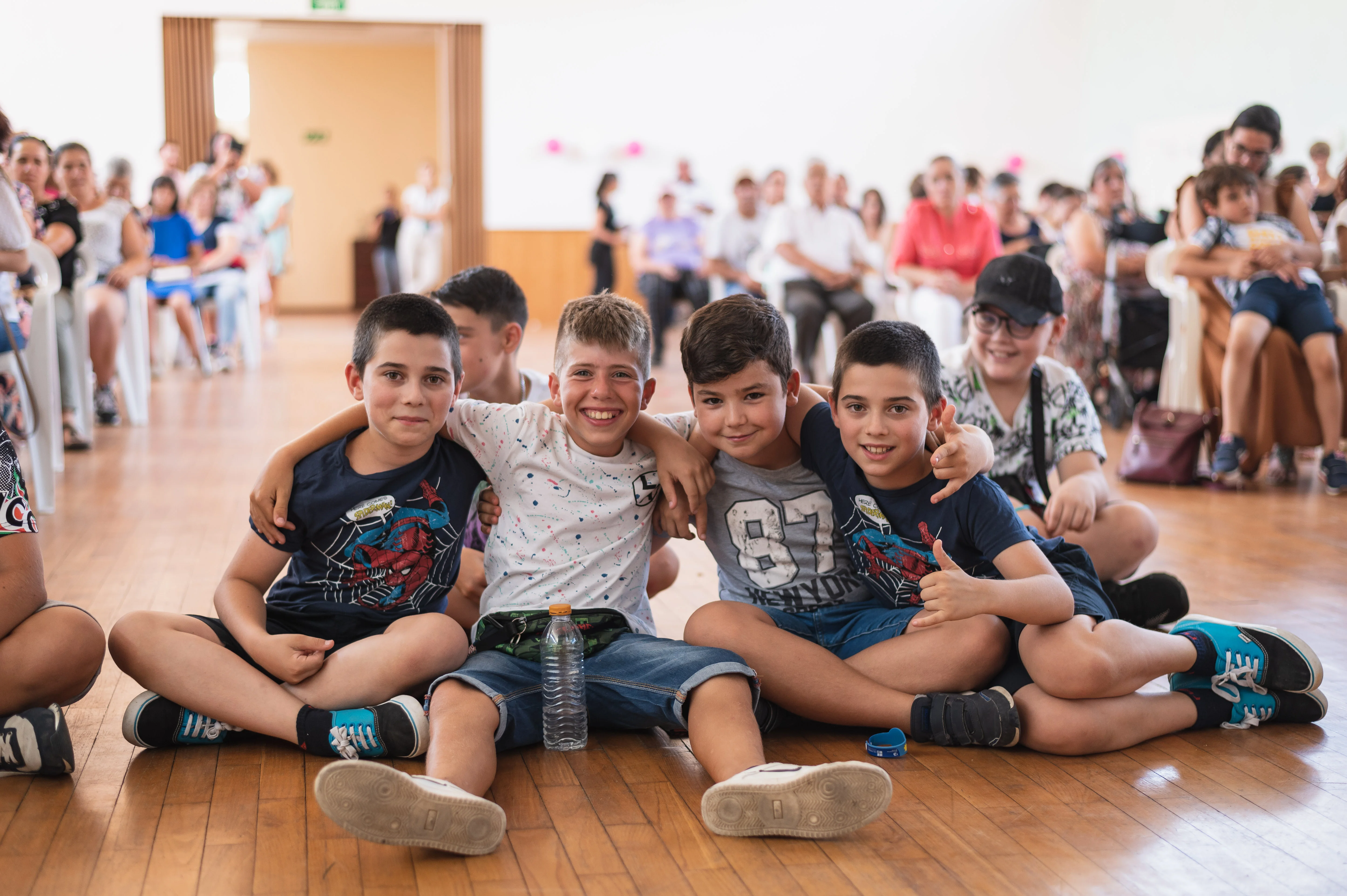 Grupo de cinco rapazes sentados no chão, sorrindo e com os braços em volta uns dos outros.