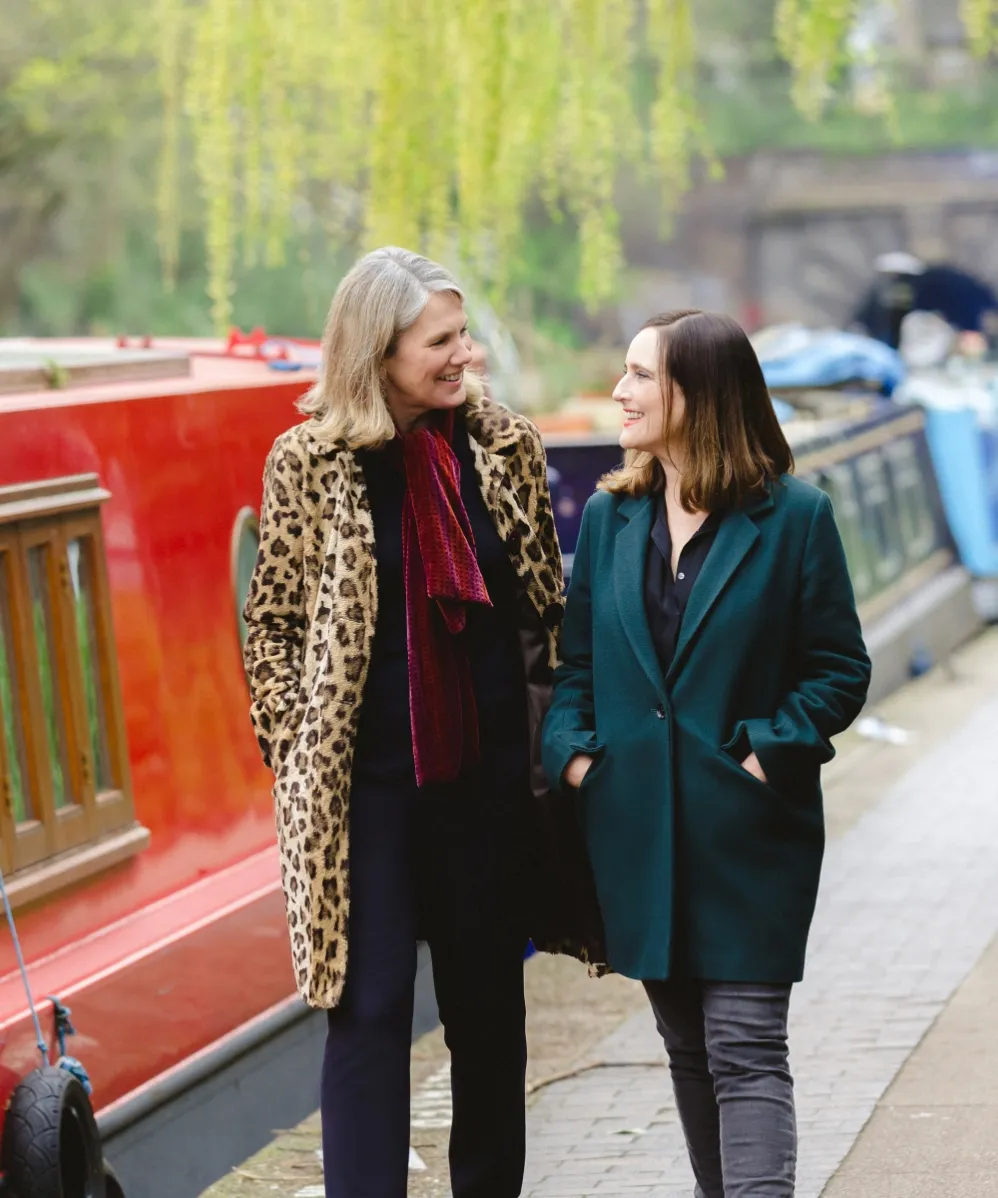 Tracey Camilleri and Samantha Rockey smiling and walking side by side along a canal with boathouses in the background.