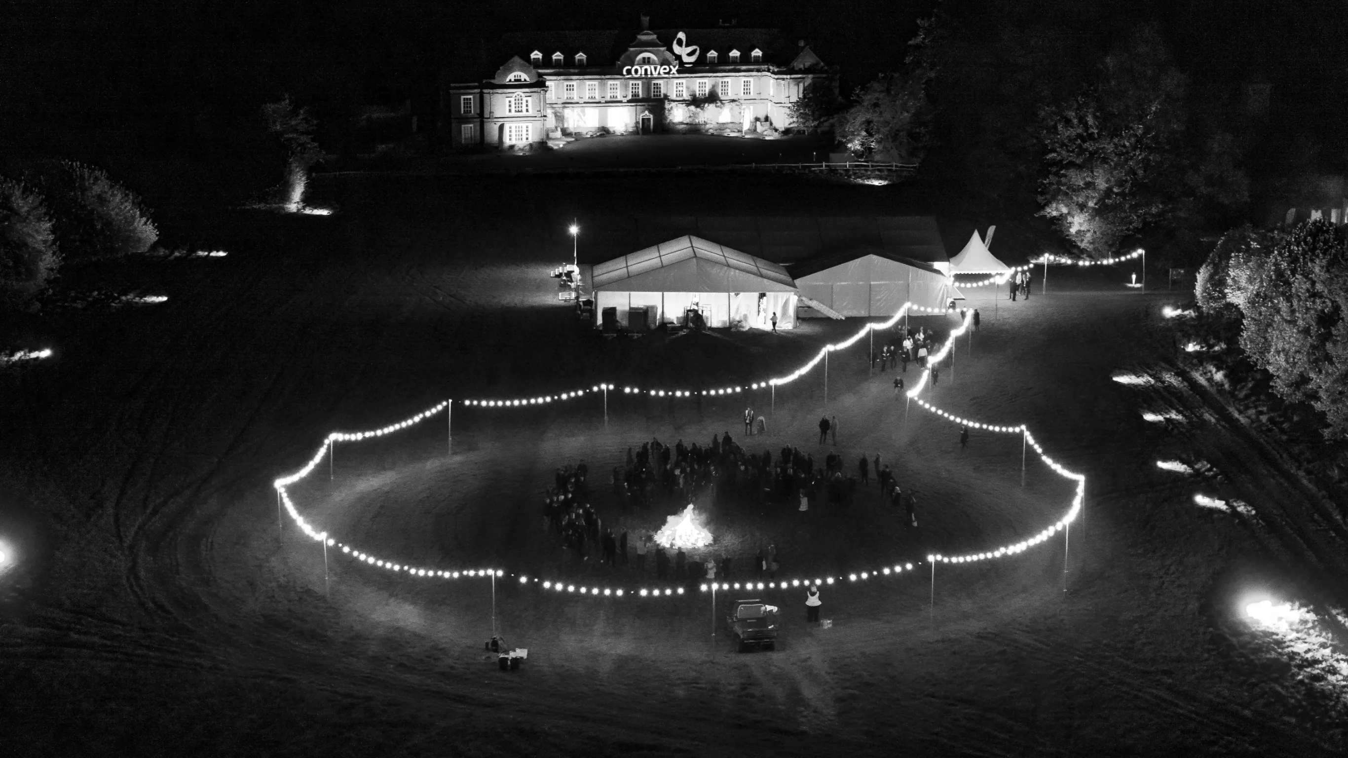 Night outdoor event with people gathered around a bonfire, string lights outlining the area and tents near a large building.