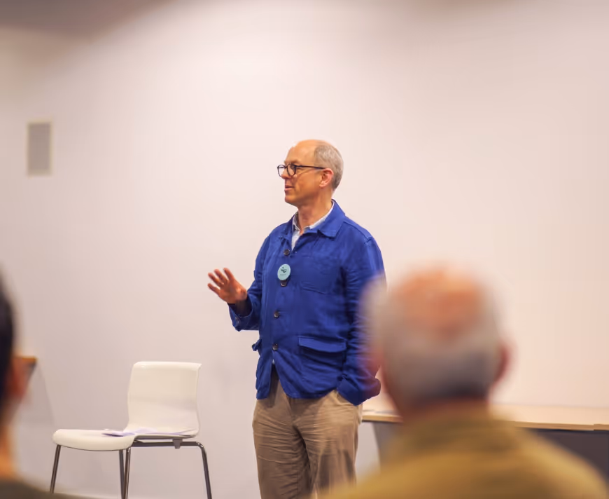 Thompson Harrison workshop participant wearing glasses and a blue jacket speaking in front of an audience in a room with white walls.