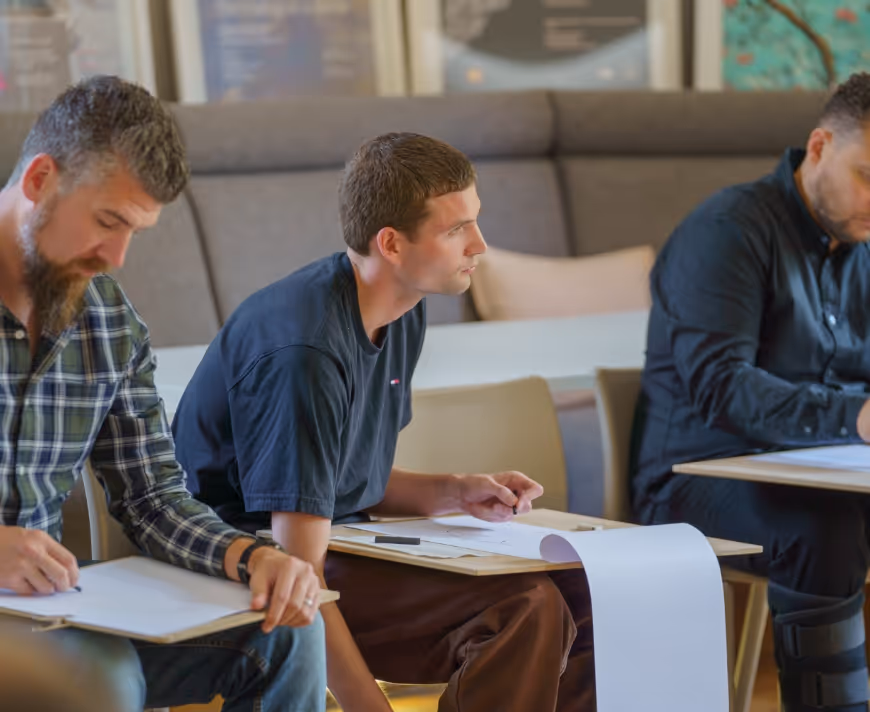 Three men sitting in a learning workshop setting, writing on paper attached to clipboards.