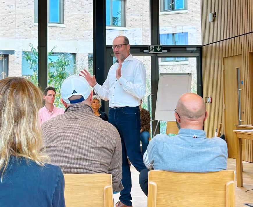 Man in white shirt gesturing while speaking to a seated group in a modern room with large windows, as at Thompson Harrison team development day.