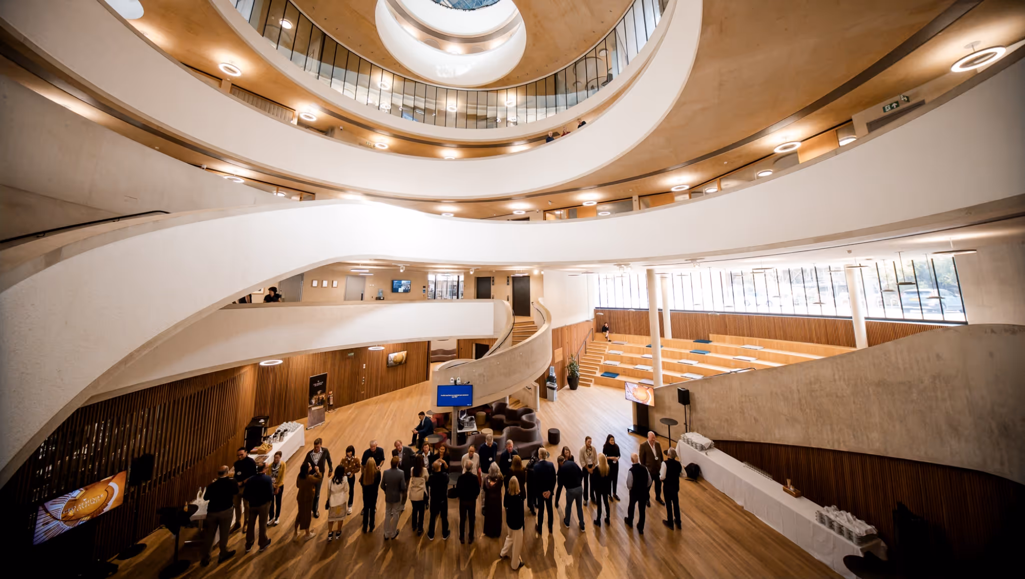 People gathered in a modern multi-level atrium with curved balconies and wooden floors.