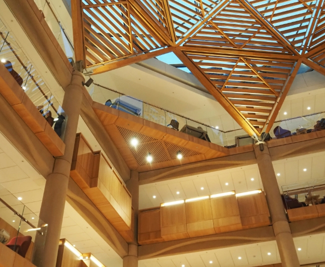 Interior view of a modern multi-level building with wooden accents and a geometric wooden ceiling structure.