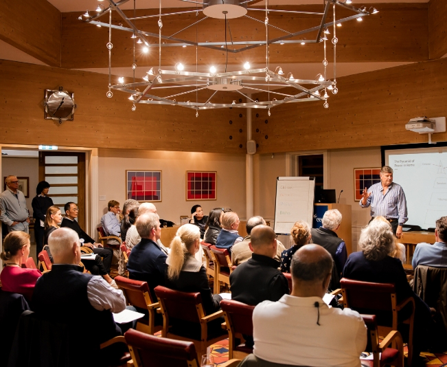 Man giving a presentation to a seated audience in a wood-paneled room with modern lighting.
