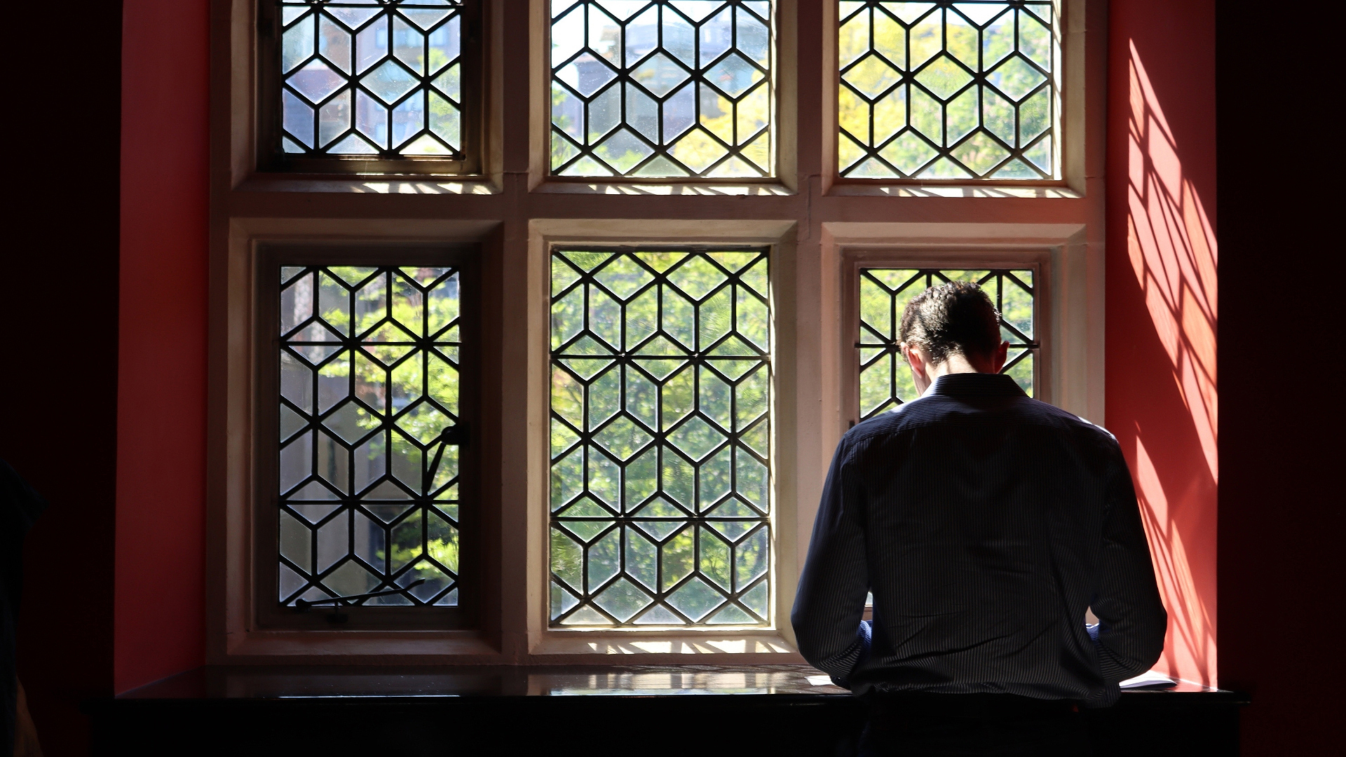 A man with glasses and a dark shirt in a Thompson Harrison website, standing by a decorative window with geometric panes and sunlight illuminating the room.