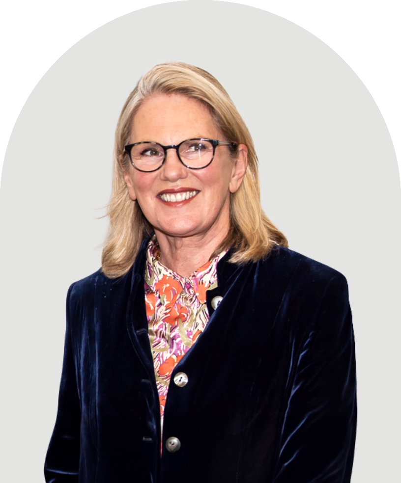 Smiling Tracey Camilleri  wearing glasses, a floral shirt, and a dark velvet jacket against a light background.