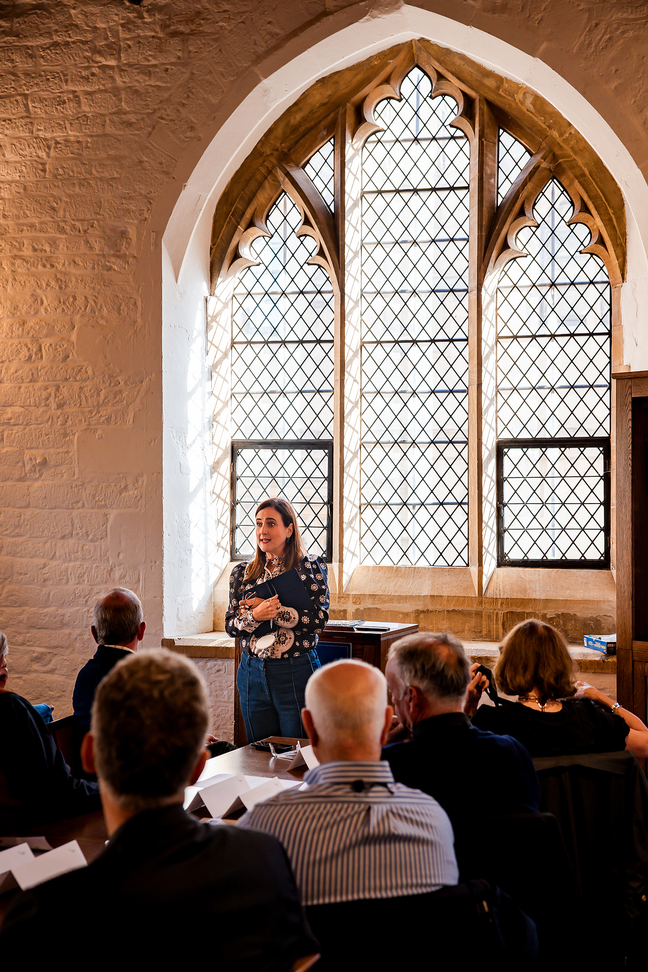 Woman speaking in front of an audience inside a room with a large arched window.