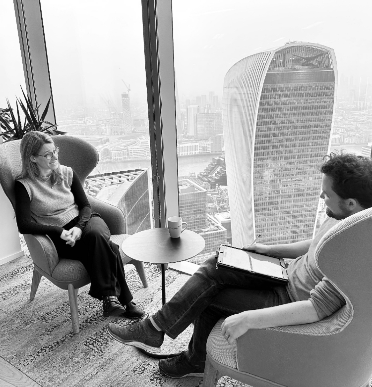 Two people sitting in armchairs by a window with a city view, one holding a tablet and a coffee mug on a small table.