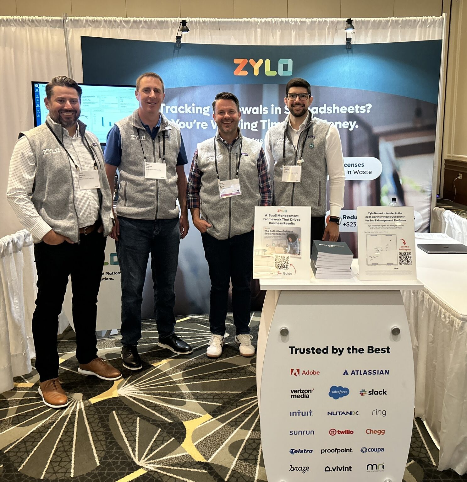 Four men wearing Zylo vests standing at a Zylo booth with a banner and table showing business and partner logos.