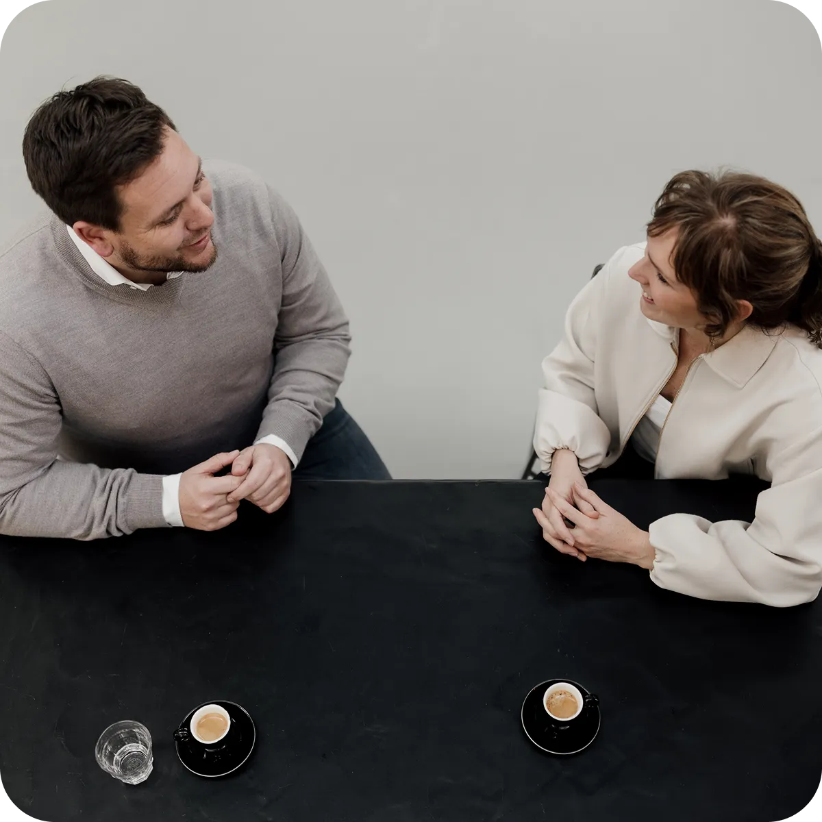 Man and woman sitting across from each other at a black table, each with an espresso cup, smiling and talking.