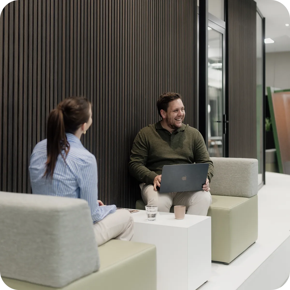 Man and woman sitting on modern chairs in an office, the man smiling and holding a laptop.
