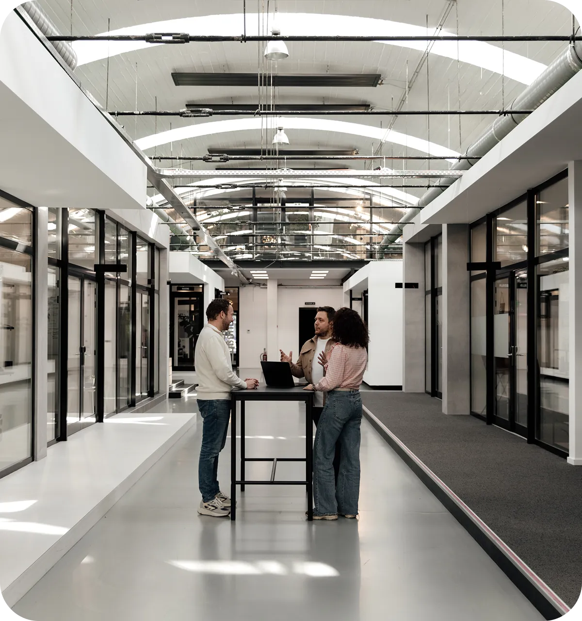 Three people standing and having a discussion around a high table with a laptop in a modern office hallway.
