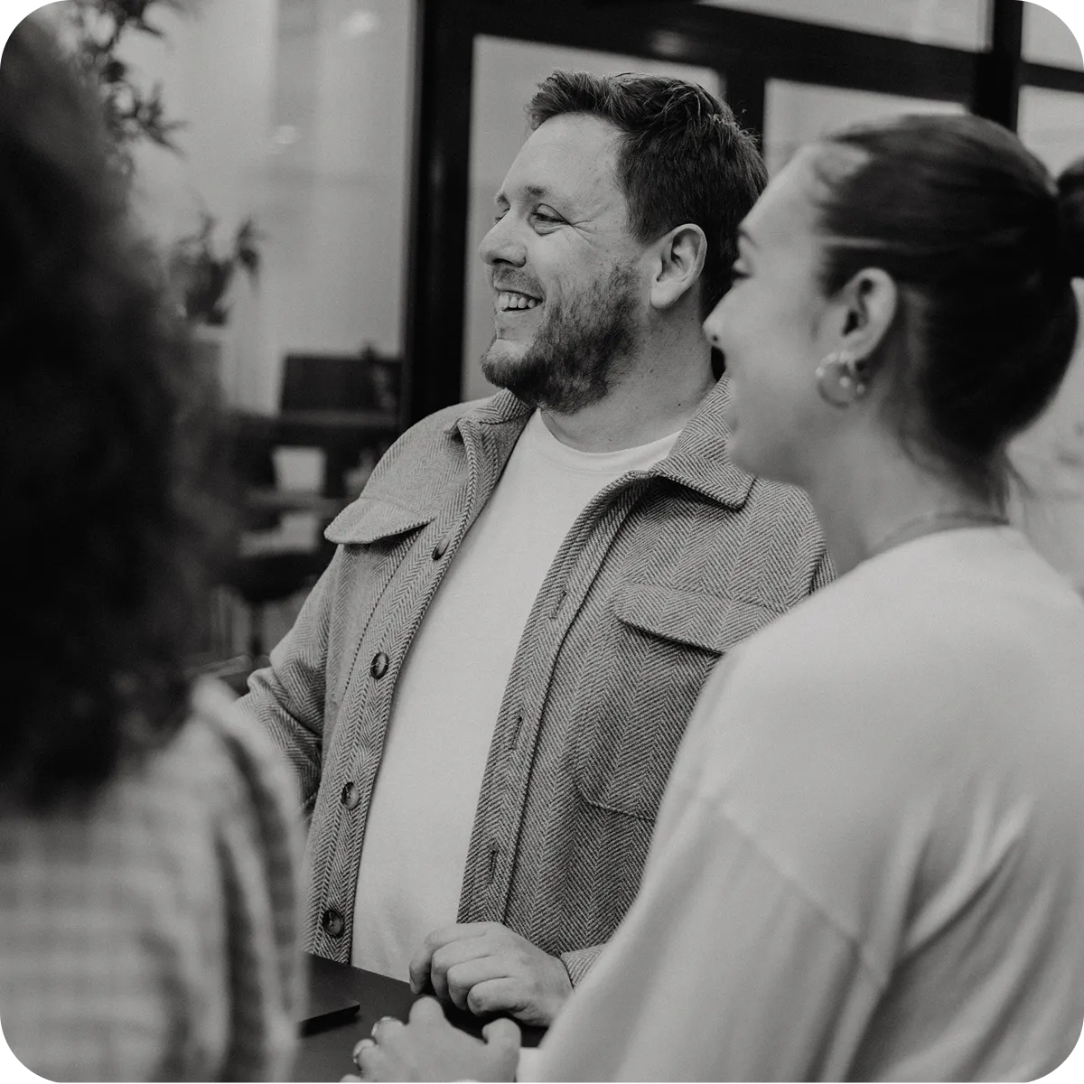 Three people smiling and interacting in a casual indoor setting, with one man facing left and two women partially visible.