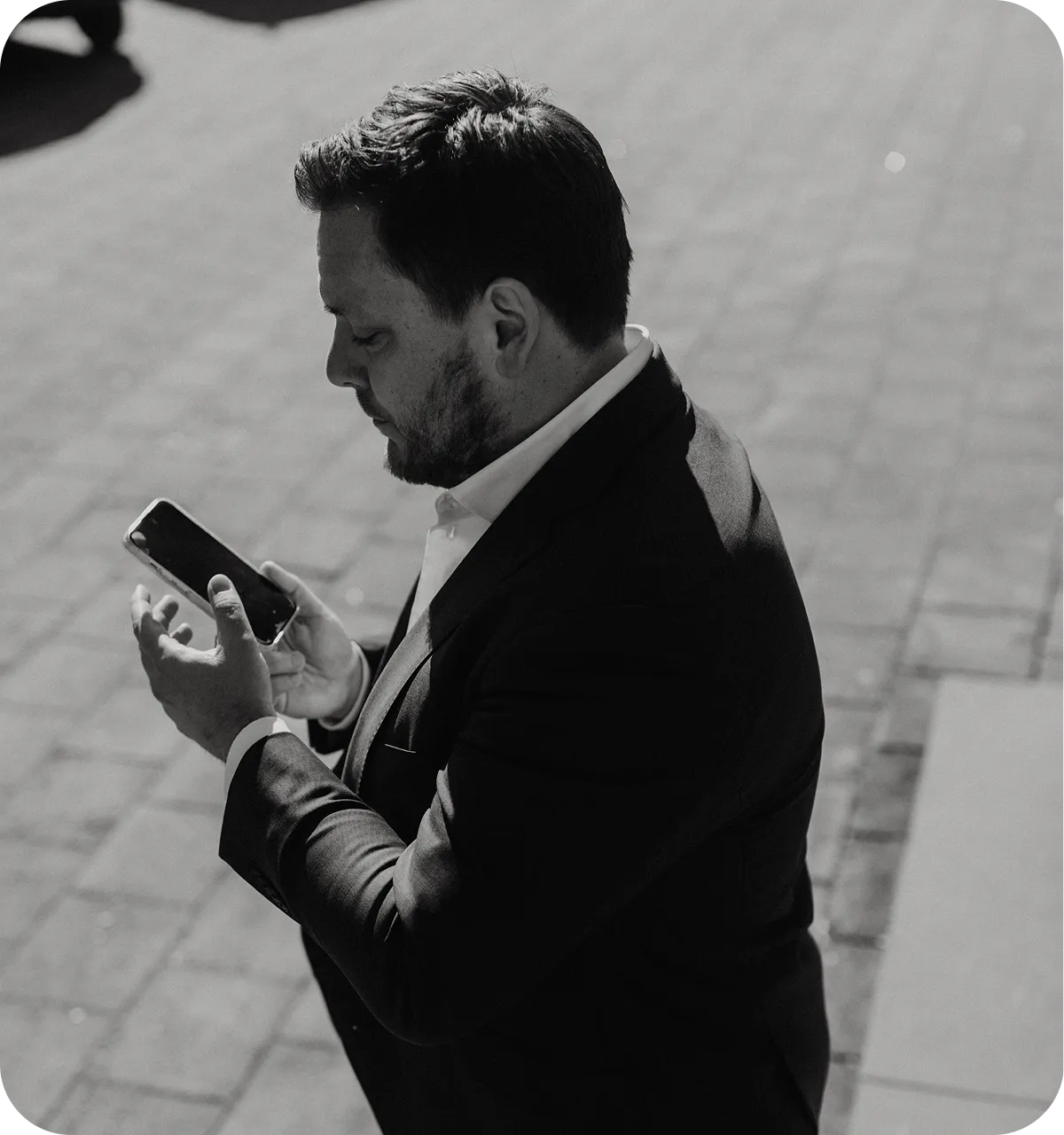 Man in a suit looking at a smartphone while standing on a paved outdoor area.