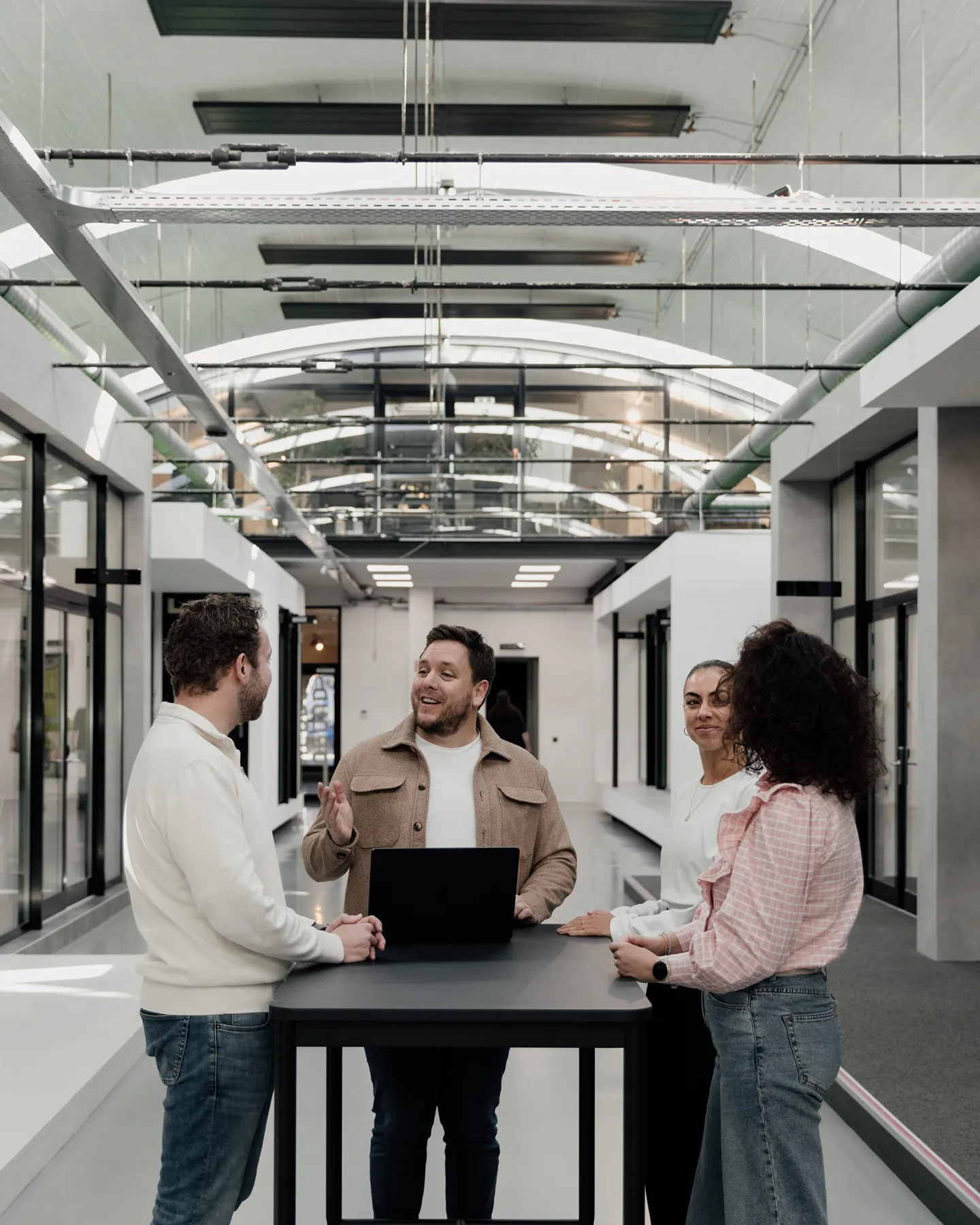 Three people standing around a high table with a laptop in a modern open office space with glass doors.