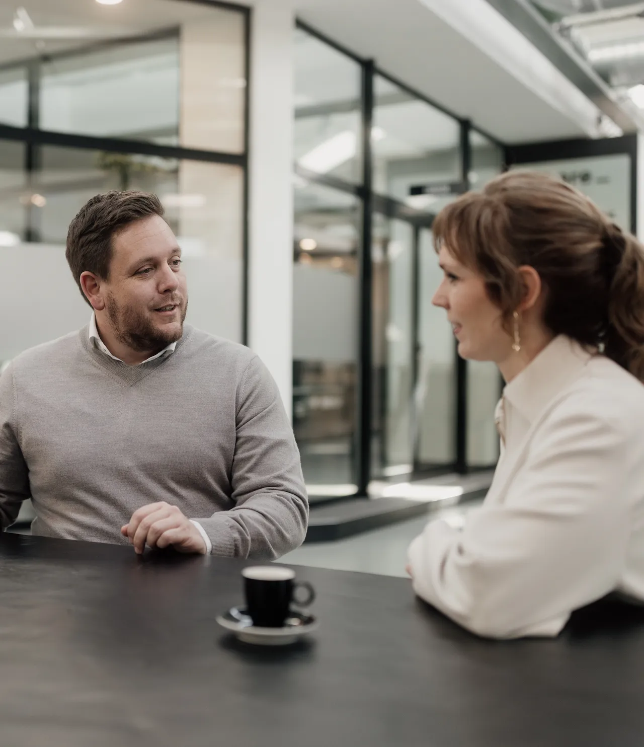 Three people standing and discussing around a high table with a laptop in a modern office corridor.