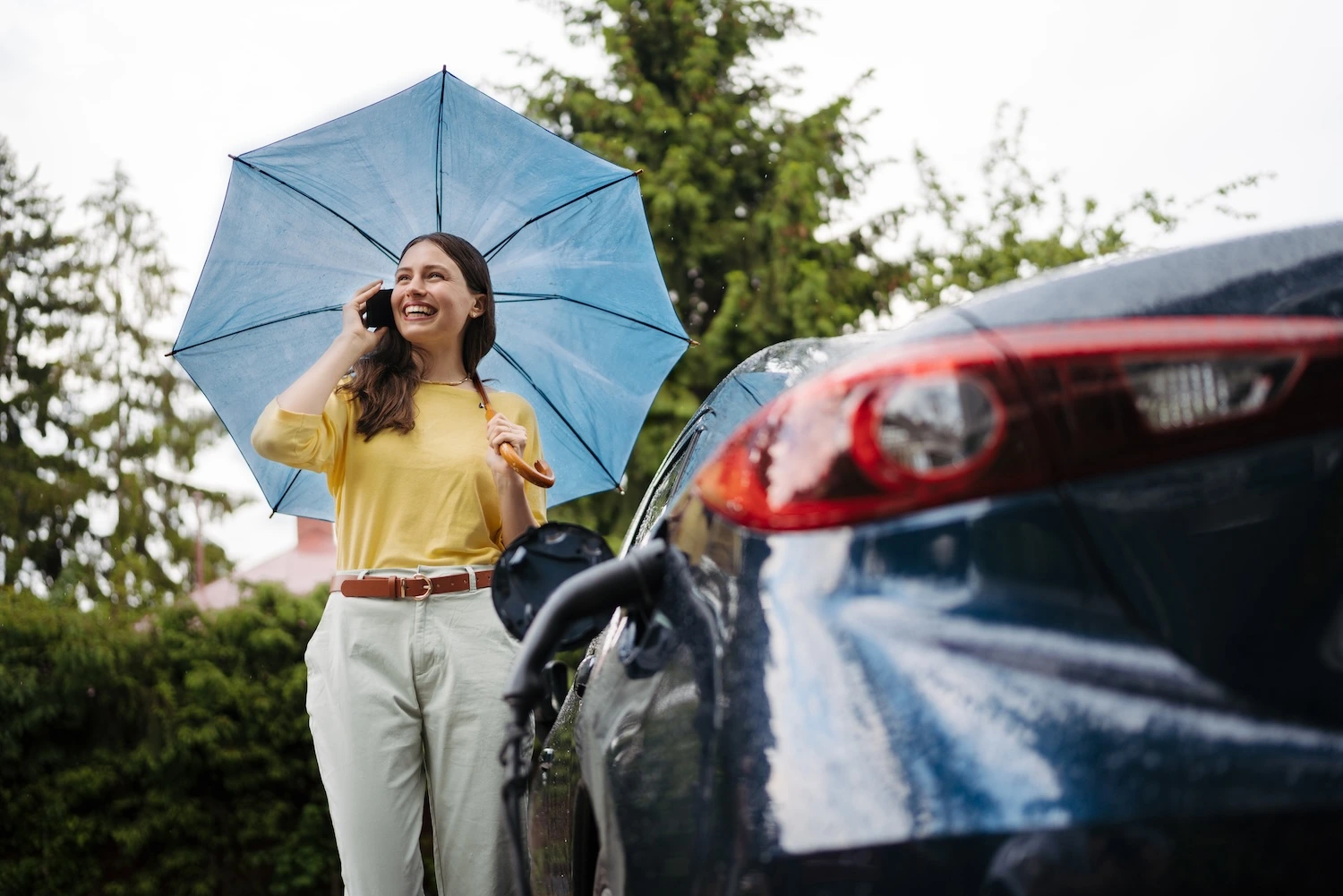 Smiling woman on the phone holding an umbrella as she approaches the driver-side door of a blue car.