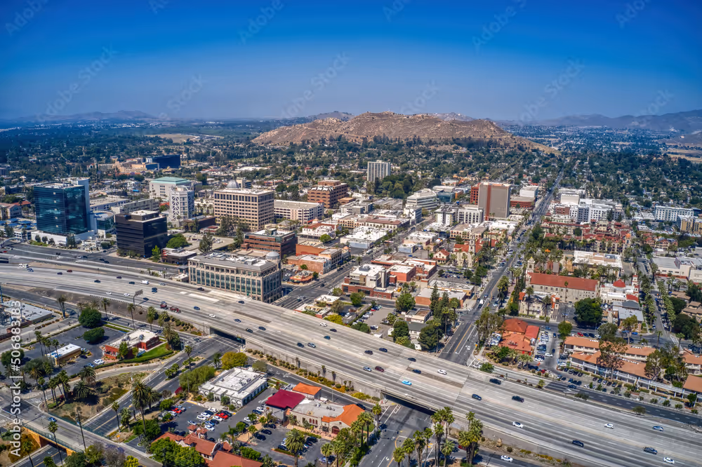 Aerial view of a cityscape with highways, buildings, and hills in the background under a clear blue sky.