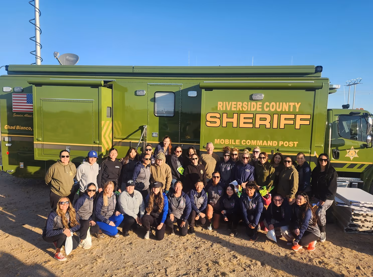 Group of people posing in front of a green Riverside County Sheriff Mobile Command Post truck on a sunny day.