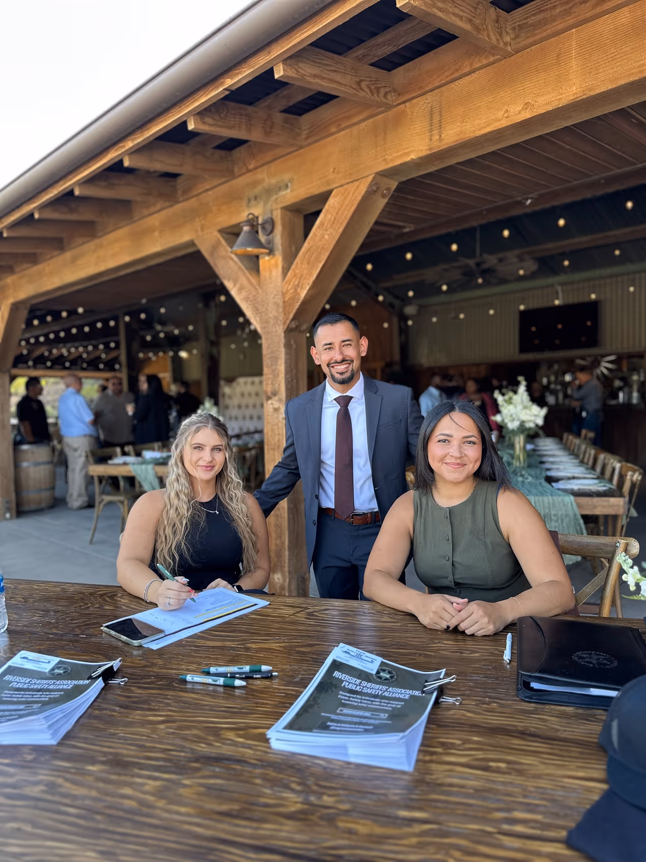 Three smiling people posing at a wooden table with documents and pens under a wooden pavilion at an outdoor event.