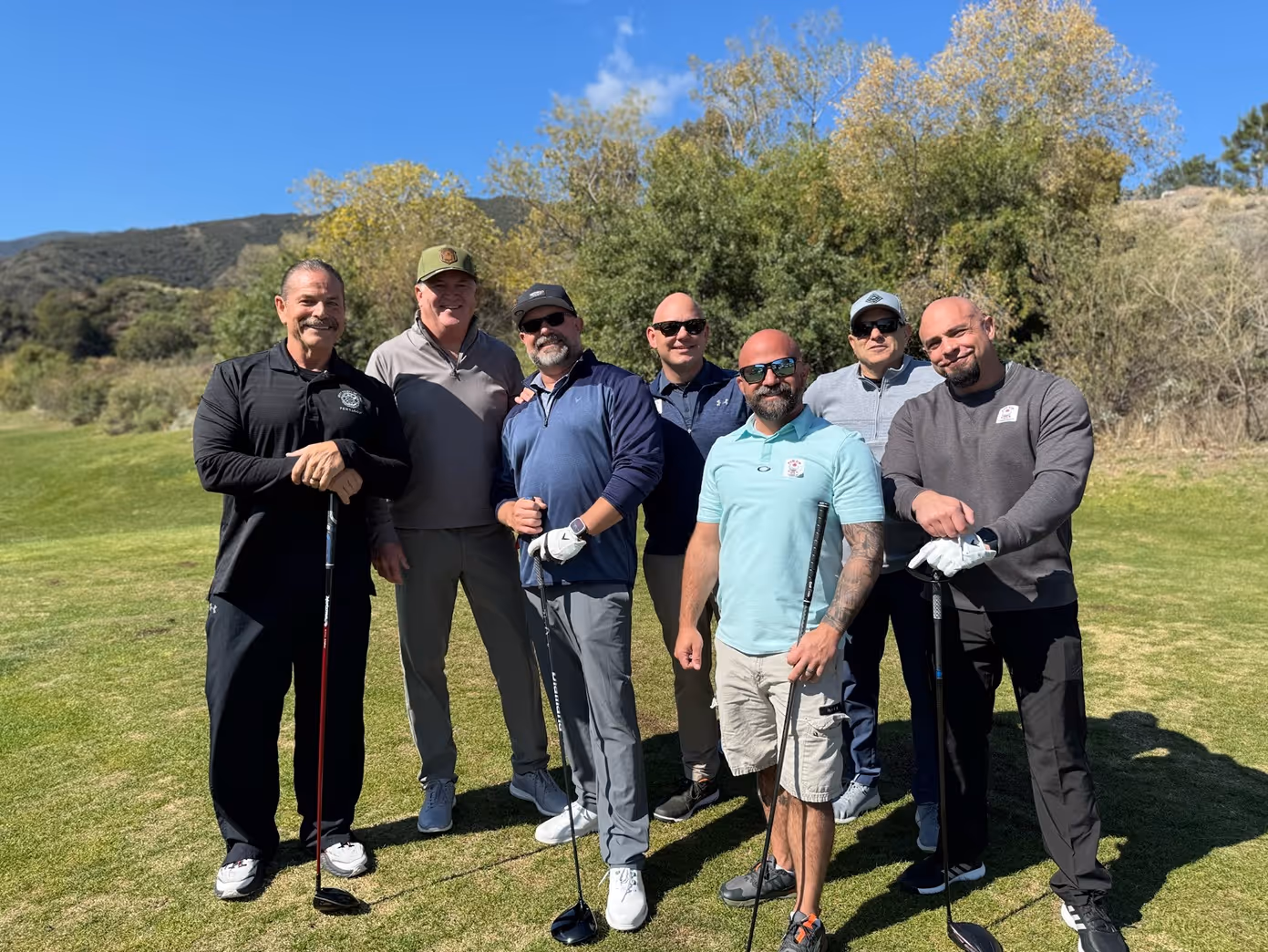 Group of seven men standing together on a golf course holding golf clubs on a sunny day.