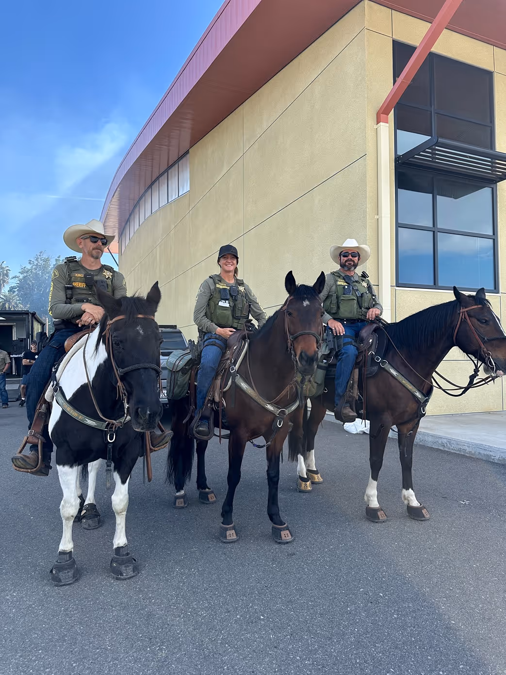 Three sheriff officers wearing uniforms and hats riding horses in front of a tan building on a paved surface.
