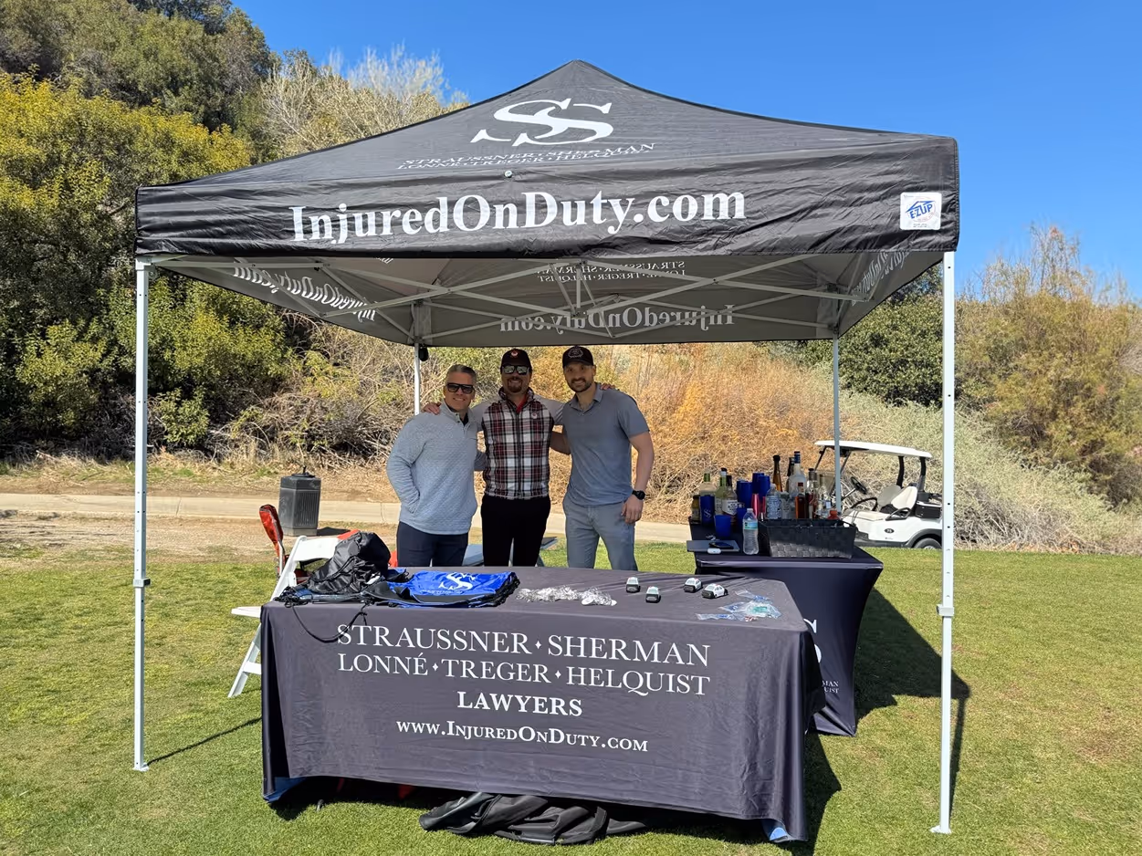 Three men standing under a black canopy tent with 'InjuredOnDuty.com' and 'Straussner Sherman Lonne Treger Helquist Lawyers' branding, outdoors on grass with trees in the background.