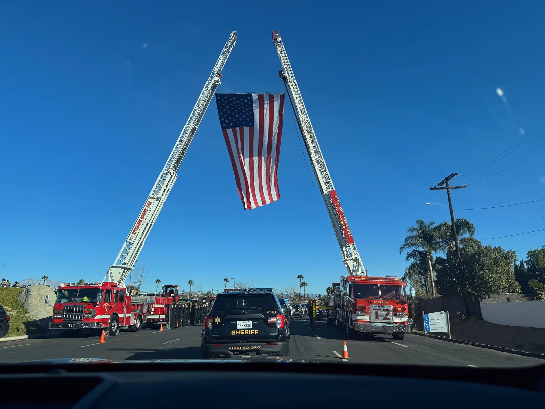 Two fire truck ladders extended upward holding a large American flag over a street with a sheriff SUV and a line of firefighters below.