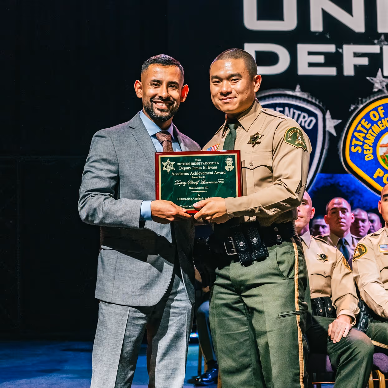 Two men smiling while holding a Riverside Sheriff's Association Deputy James B. Evans Academic Achievement Award plaque on stage with other deputies seated in the background.