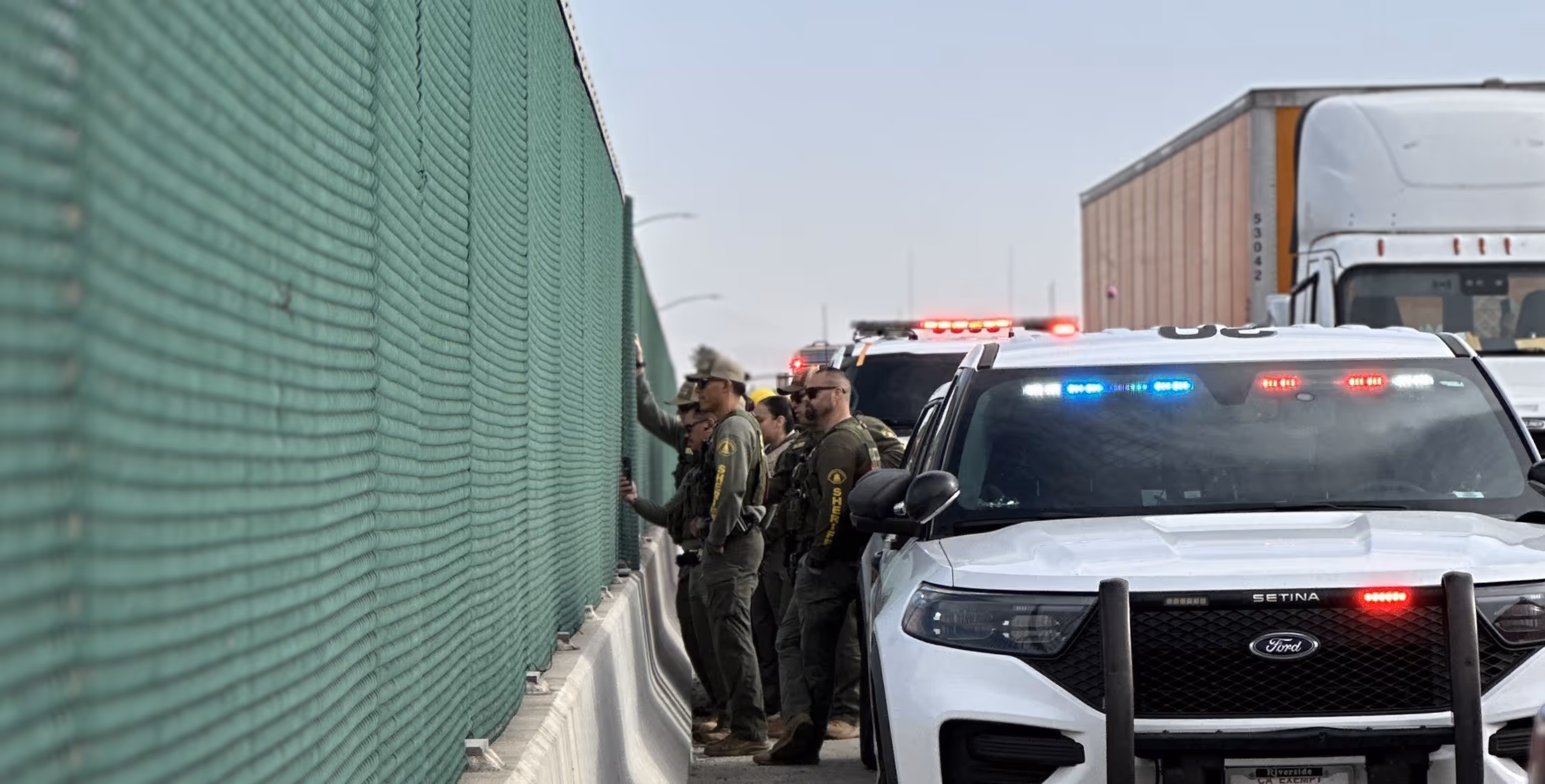 Group of sheriff officers standing by a chain-link fence next to police vehicles with flashing lights on a highway.