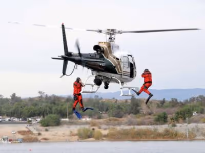 Two Riverside County Sheriff's deputies in orange drysuits rappelling from a helicopter over a lake during a water rescue training exercise.