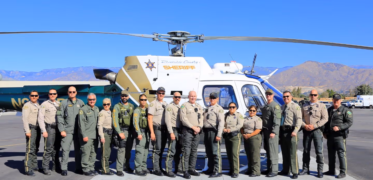 Group of Riverside County Sheriff's deputies standing in front of a sheriff helicopter with mountains in the background.