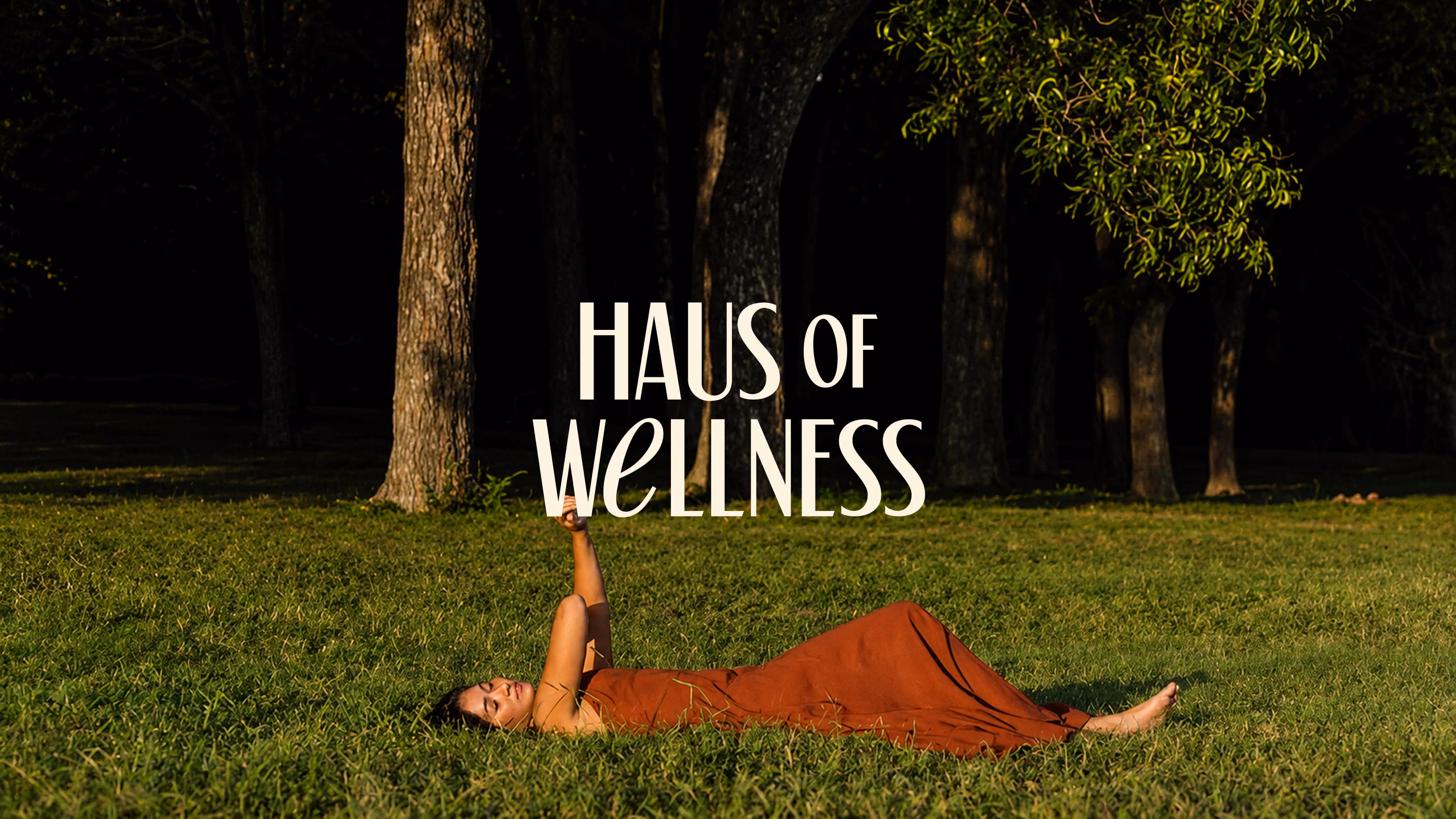 Woman in a long orange dress lying on green grass in a sunny park with trees, with the text 'HAUS OF WELLNESS' above her.