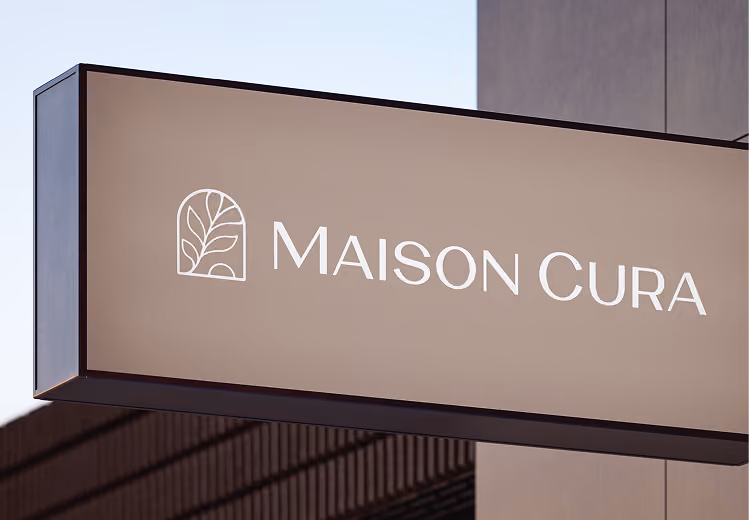 Outdoor sign with a stylized leaf logo and the text 'MAISON CURA' on a beige background.