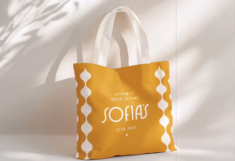 Yellow tote bag with white wavy vertical stripes on the sides and white handles, featuring the text 'Authentic Greek Cuisine SOFIA'S ESTD 2025' in white centered on the front.