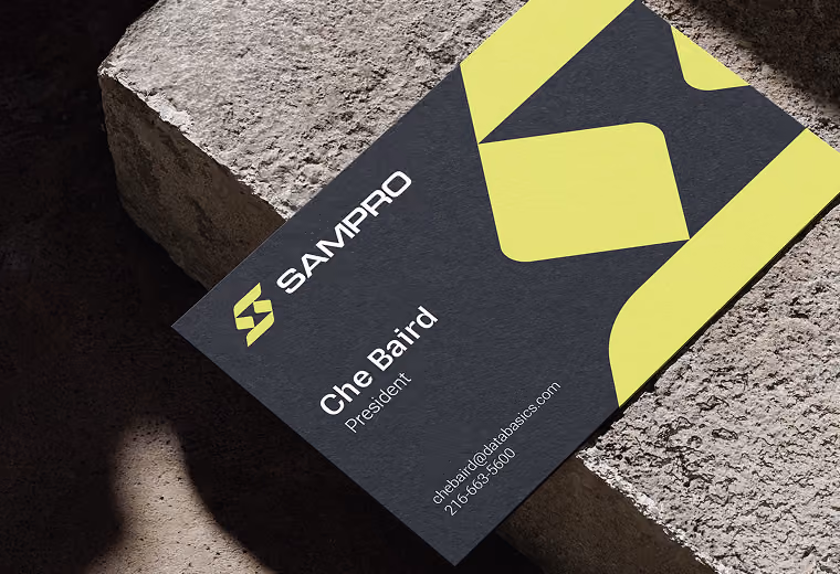 Black business card on textured stone surface with bright yellow geometric design, showing company name SamPro, individual named Che Baird, President, email chebaird@databasics.com, and phone number 216-663-5600.