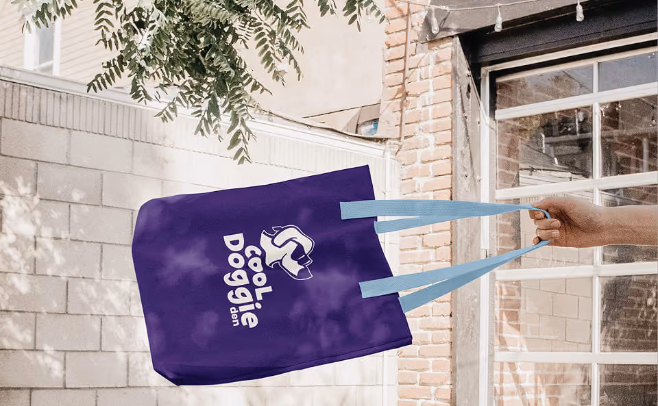 A hand holding a purple tote bag with light blue handles, displaying a white logo and text that reads 'Cool doggie den' featuring a dog illustration, against a backdrop of a brick and white wall with greenery above.